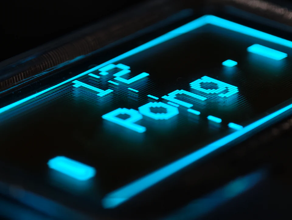 Macro photograph of Pong on the transparent OLED, with cyan score digits, ball, paddles, and individual pixel cells visible.